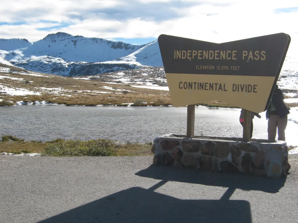 Sign for Independence Pass, Continental Divide, Elevation 12,095 feet. Snow capped mountains in the background with a small pond. 